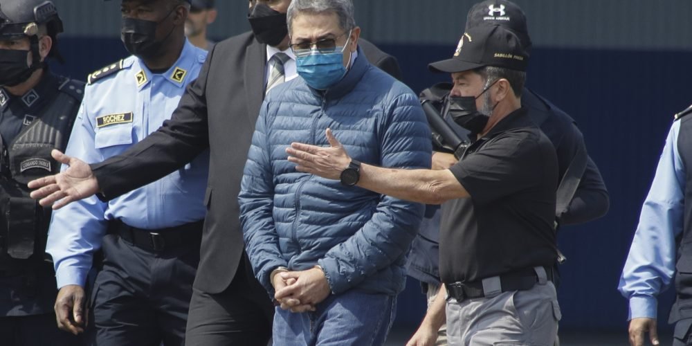 ARCHIVO - El expresidente hondureño Juan Orlando Hernández, en el centro, es llevado esposado a un avión que lo espera mientras es extraditado a Estados Unidos para enfrentar cargos de tráfico de drogas en una base de la Fuerza Aérea en Tegucigalpa, Honduras, el 21 de abril de 2022. El juicio de Hernández en Nueva York comienza el lunes 12 de febrero de 2024. (AP Foto/Elmer Martinez, Archivo)