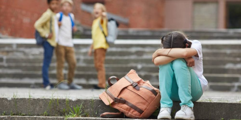 Full length portrait of crying schoolgirl sitting on stairs outdoors with group of teasing children bullying her in background, copy space